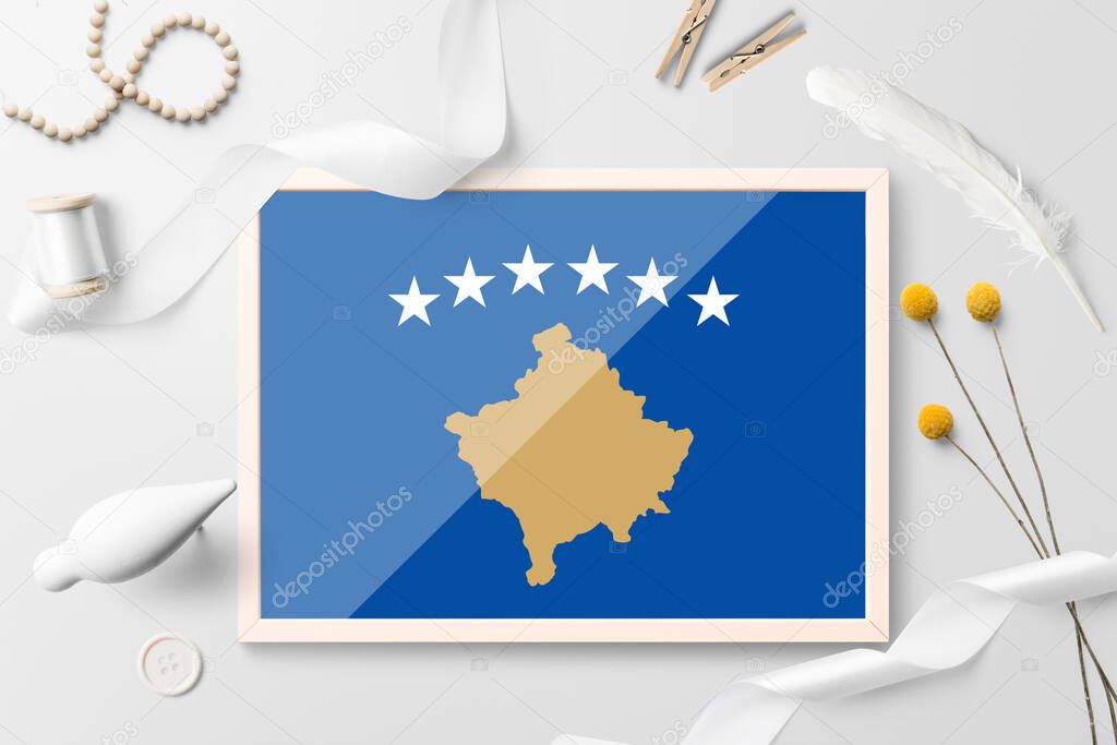 Kosovo flag in wooden frame on white creative background. White theme, feather, daisy, button, ribbon objects.