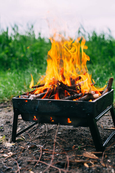A small folding portable brazier filled with burning wood. Cooking on the grill. Safe fire in nature.