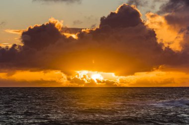Gün batımında bulutlarla birlikte gökyüzünün altındaki sonsuz dalgalı deniz yüzeyinin resimsel görüntüsü.