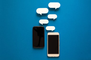 Kağıt konuşma balonları üzerinde mavi arka plan ile akıllı telefonlar. İletişim kavramı. Üstten Görünüm. Uzay kopyalayın. Kağıt kompozisyon