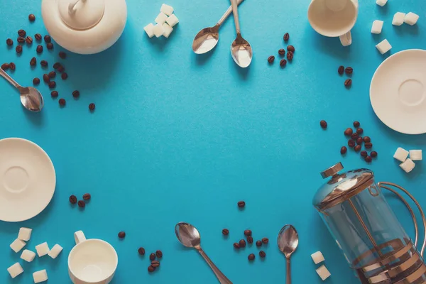 Various coffee making accessories: French coffee press, cups, saucers, coffee beans, spoons and sugar on blue paper background. Good morning concept. Space for copy. Top view. Flat lay. Toned