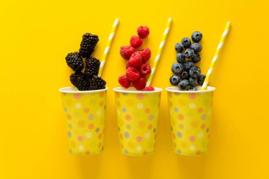 Kağıt bardak taze raspbery, yaban mersini ve blackberry sarı Kağıt arka plan üzerinde yalan. Üstten görünüm. Düz yatıyordu. Uzay kopyalayın. Yaz içki kavramı. Limonata için malzemeler