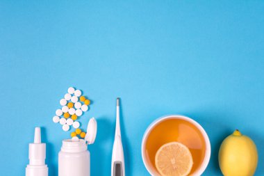 Tıbbi elektronik termometre, hap, burun için sprey ilaç şişesiyle ve mavi arka plan üzerinde limonlu çay. Sağlık kavramı. Bitkisel ve kimyasal ilaç. Üstten görünüm. Uzay kopyalayın. Düz yatıyordu