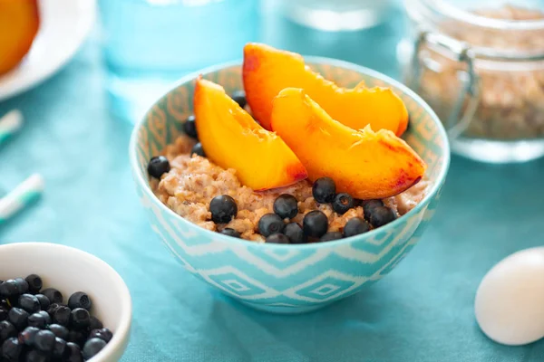 Porridge bowl oatmeal with peach and blueberries on turquoise background. Soft focus. Healthy eating concept.