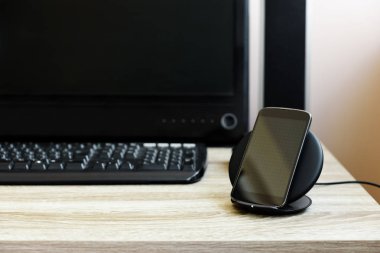 Siyah akıllı telefon kablosuz şarj cihazı ile bilgisayar monitörü, klavye ve hoparlörler ahşap masa üzerinde karşı şarj. Kablosuz şarj veya dijital teknoloji konsepti. Çalışma alanı