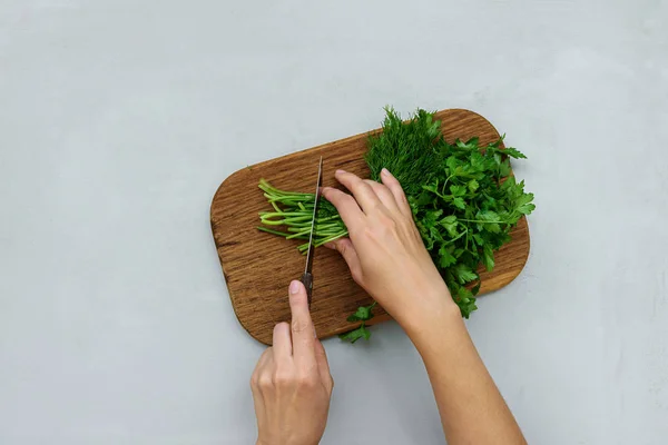 Feminine hands chopping fresh green parsley and dill or fennel on cutting boar on gray wooden table. Top view. Copy space. Harvesting concept