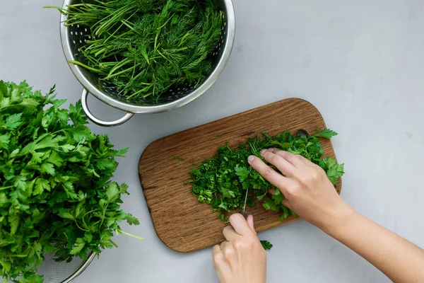 Feminine hands chopping fresh green parsley and dill or fennel on cutting boar on gray wooden table. Top view. Copy space. Harvesting concept