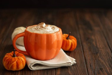 Balkabağı şeklinde bir kupada, sonbahar dekoruyla çevrili, krema ve baharatlı, kabak şeklinde bir latte.