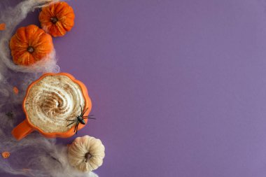 Cadılar Bayramı temalı düz yatış, balkabaklı latte, dekoratif balkabakları ve mor arka planda örümcek ağları..