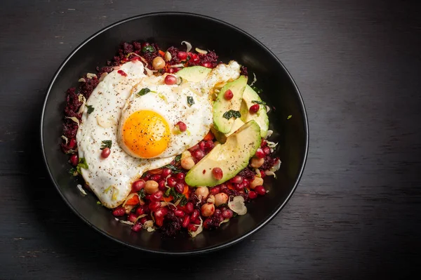 Miso veggie breakfast bowl with beets, carrots, Brussels sprouts, kale, chickpeas, and topped with pomegranate seeds, avocado and fried egg served in a dark bowl. Background is a rough, dark wood surface. Room for copy on right.