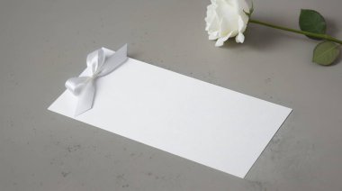 White invitation with bow on a concrete backdrop with a white rose