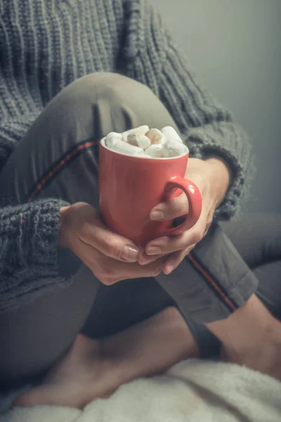 Red cup with hot cocoa and marshmallows in the hands of a woman in a warm cozy jacket close-up