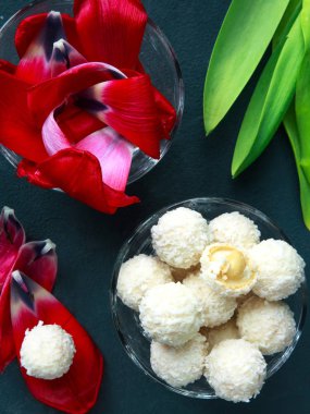 Kırmızı Terry laleleri ve bademli yuvarlak şekerler ve karanlığın yatay arka planında çiçek yaprakları üzerinde cam bir kasede hindistan cevizi. Ziyafet konsepti.