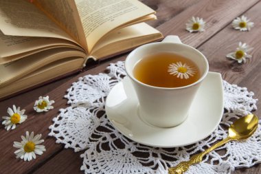 Beyaz fincanda papatya çayı ve arka planda koyu ahşap masa konsepti üzerine bir kitap. Sağlıklı bitkisel içecek ve romantik bir kahvaltı.