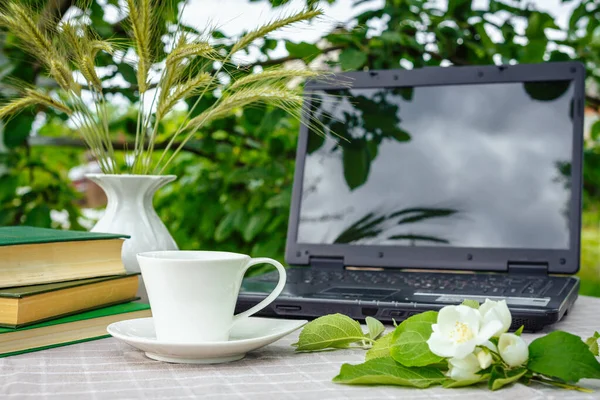 A cup with tea, a laptop, a computer, a bookshine table in the garden on a summer morning, a place to work, a concept of freelancing