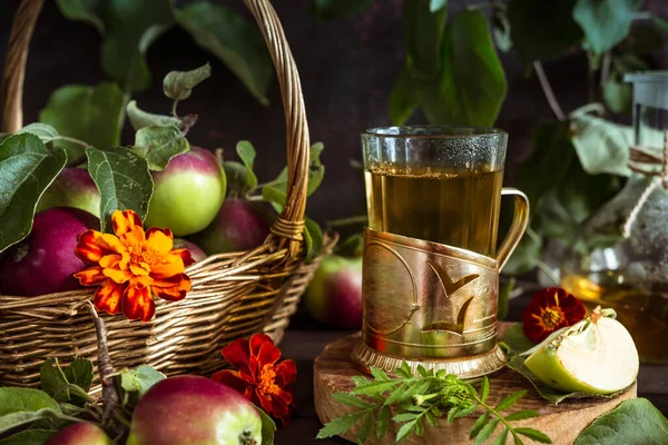 Apple tea in a glass goblet and a decanter and a basket with ripe apples on a wooden table in vintage style
