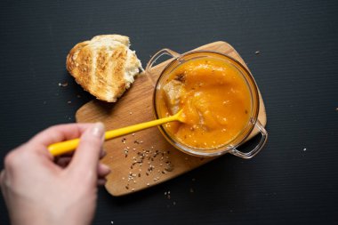 genç bir kızın eli balkabağı çorbasına batırılmış sarı bir kaşık tutuyor, siyah bir arka plan üzerinde tahta bir tahta, bir parça ekmek, susam ve keten tohumu