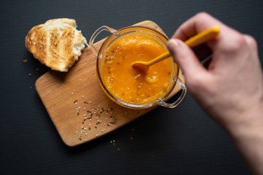 genç bir kızın eli balkabağı çorbasına batırılmış sarı bir kaşık tutuyor, siyah bir arka plan üzerinde tahta bir tahta, bir parça ekmek, susam ve keten tohumu