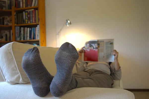 男はソファの上に足を上げて 新聞を読んでいる 少し摩耗した靴下に焦点を当てます 壁のテキスト用のスペース — ストック写真