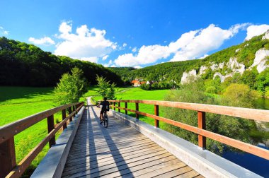A young cyclist on a wooden bridge with the typical landscape of the upper danube valley in the background clipart