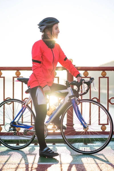 Portrait of Young Smiling Female Cyclist in Bright Orange Jacket Resting with Road Bicycle in the Cold Sunny Autumn Day. Healthy Lifestyle Concept.
