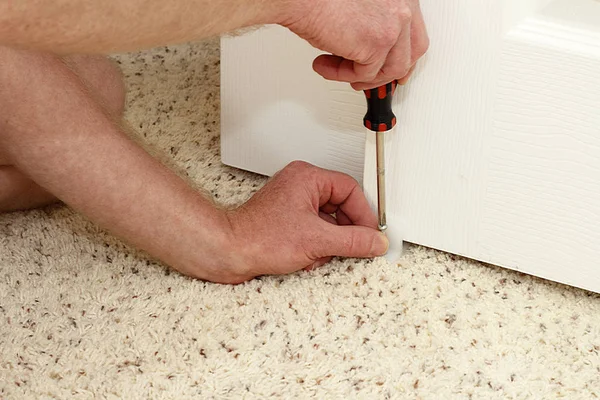 Caucasian hands of an adult male using a screwdriver to secure a white plastic closet door guide into a carpeted floor of a home. Securing a closet door floor guide