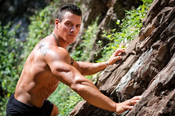 Handsome, muscular, shirless climber, looking in camera and climbing stone wall