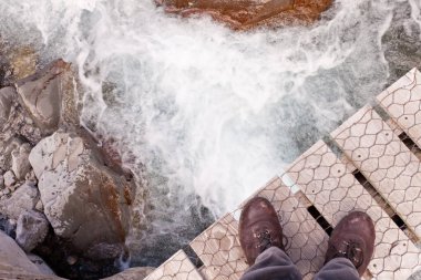 Booted feet of man standing at the edge of a narrow wooden bridge over clear fresh white water rushing through a rocky gorge clipart
