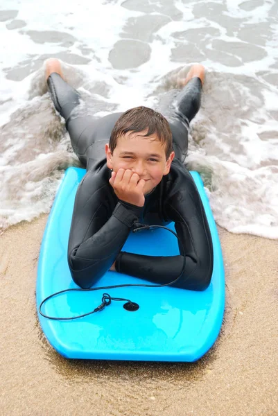 微笑的青少年冲浪者穿着潜水衣躺在他的冲浪板在海滩上 — 图库照片