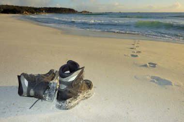 A pair of boots on a sandy beach in the early light of dawn Footprints lead into the sea but do not come out again. Space for copy on the sand. clipart