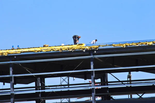 Two men are tethered to the roof of a high-story building. They are working on the roof of this new building under construction.