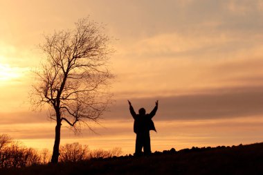 Silhouette of a man on a hill standing by a tree with his arms lifted in the air clipart