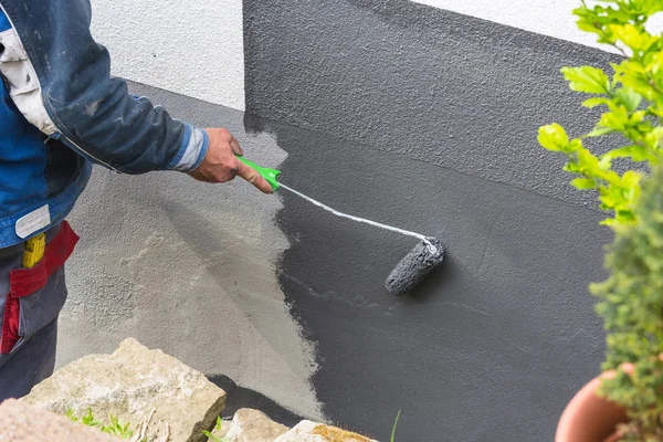 Close-up, arm of a house painter with paint roller in hand painting a house wall with gray paint.