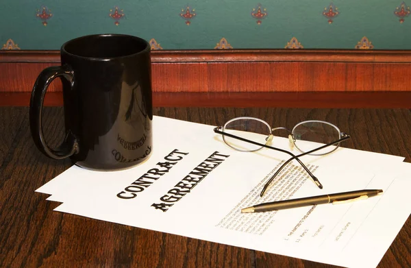 Contract agreement papers are in the decision process as shown by black coffee cup, reading glasses, and pen