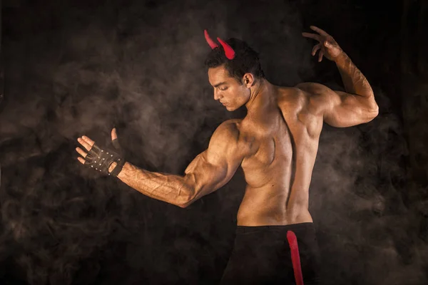 Shirtless muscular male bodybuilder dressed with devil costume on dark background