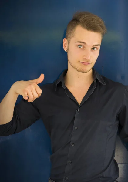 Attractive blond, blue eyed young man smiling and doing thumb up sign with one hand. Blue background