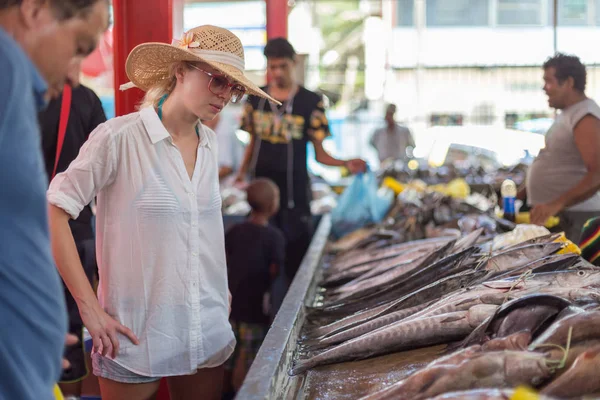 Female traveler wearing elegant colonial style white tunic and hat visiting traditional Victoria fish market on Seychelles islands.