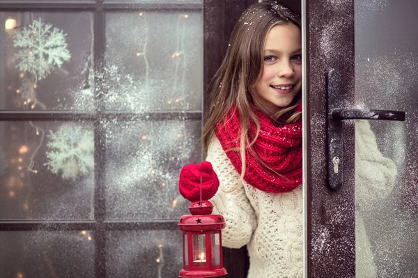 Child girl welcome guests and holding holiday lantern, snow weather, house is decorated before Christmas