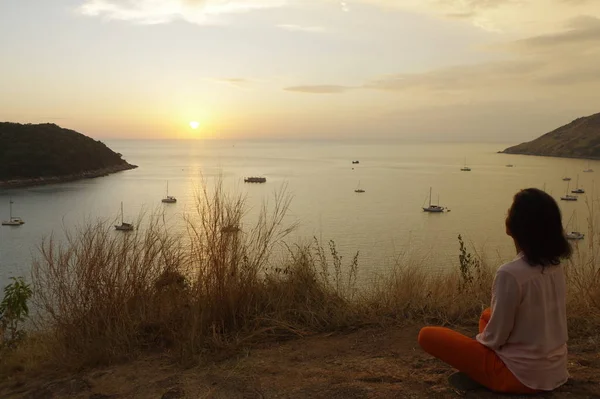 Young girl sitting in yoga lotus meditation position front to seaside on the rocks an watching the sun goes down in a golden hour. sunset over sea.
