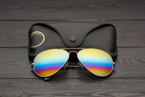 aviators sunglasses with mirrored color lenses made of glass in a gold metal frame covered with gold matte dusting on a black wooden background or table with a black leather case with a clip fastener with a gold circle and an inscription on a leather