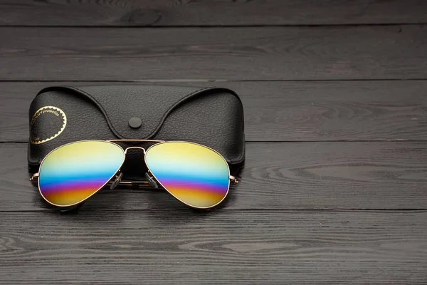 aviators sunglasses with mirrored color lenses made of glass in a gold metal frame covered with gold matte dusting on a black wooden background or table with a black leather case with a clip fastener with a gold circle and an inscription on a leather