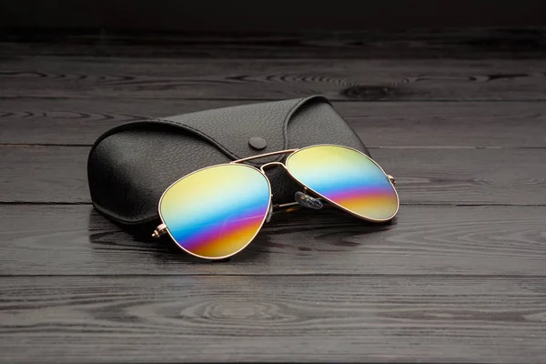 aviators sunglasses with mirrored color lenses made of glass in a gold metal frame covered with gold matte dusting on a black wooden background or table with a black leather case with a clip fastener with a gold circle and an inscription on a leather