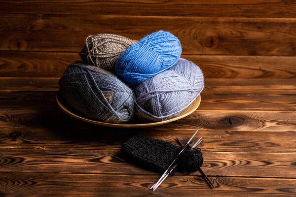 balls of gray and blue threads in a wooden plate and knitting needles with knitting on a wooden background