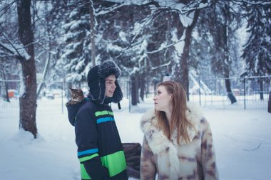 Kürk şapkalı genç bir adam, kulağı kulaklı, kapüşonlu bir kedi yavrusu ve bej kürklü bir kız kış ormanının arka planında dalgalı saçları var.