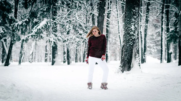 The girl dressed in a maroon sweater and white pants stands against the tree trunk against a backdrop of snow-covered winter forest — Stock Photo, Image