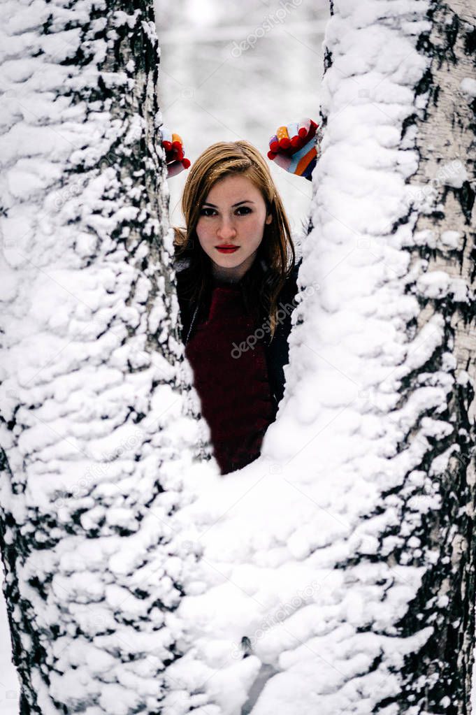girl in Burgundy jacket peeks out from behind the snowcovered bifurcated trunk of a birch. Portrait, close-up