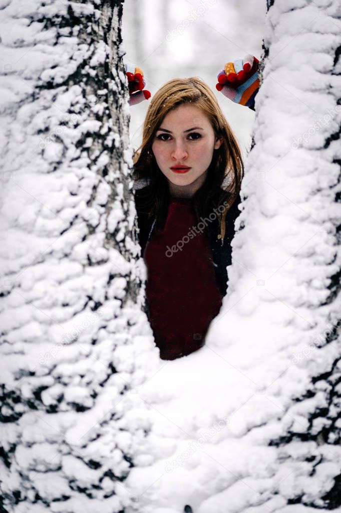 girl in Burgundy jacket peeks out from behind the snowcovered bifurcated trunk of a birch. Portrait, close-up