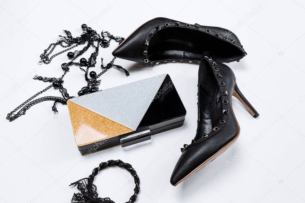 pair of black shoes decorated with metal accents, jewelery with black lace and beads and a tricolor clutch with sparkles on a white background