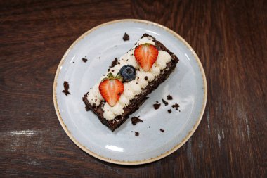 koyu ahşap arka plan üzerinde bir tabakta krem şanti, çilek ve yaban mersini ile süslenmiş brownie