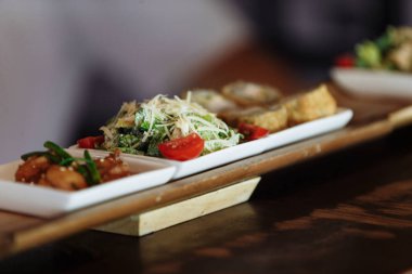 Masanın arka planında ahşap standlar üzerinde beyaz kare tabaklarda çeşitli Japon yemekleri